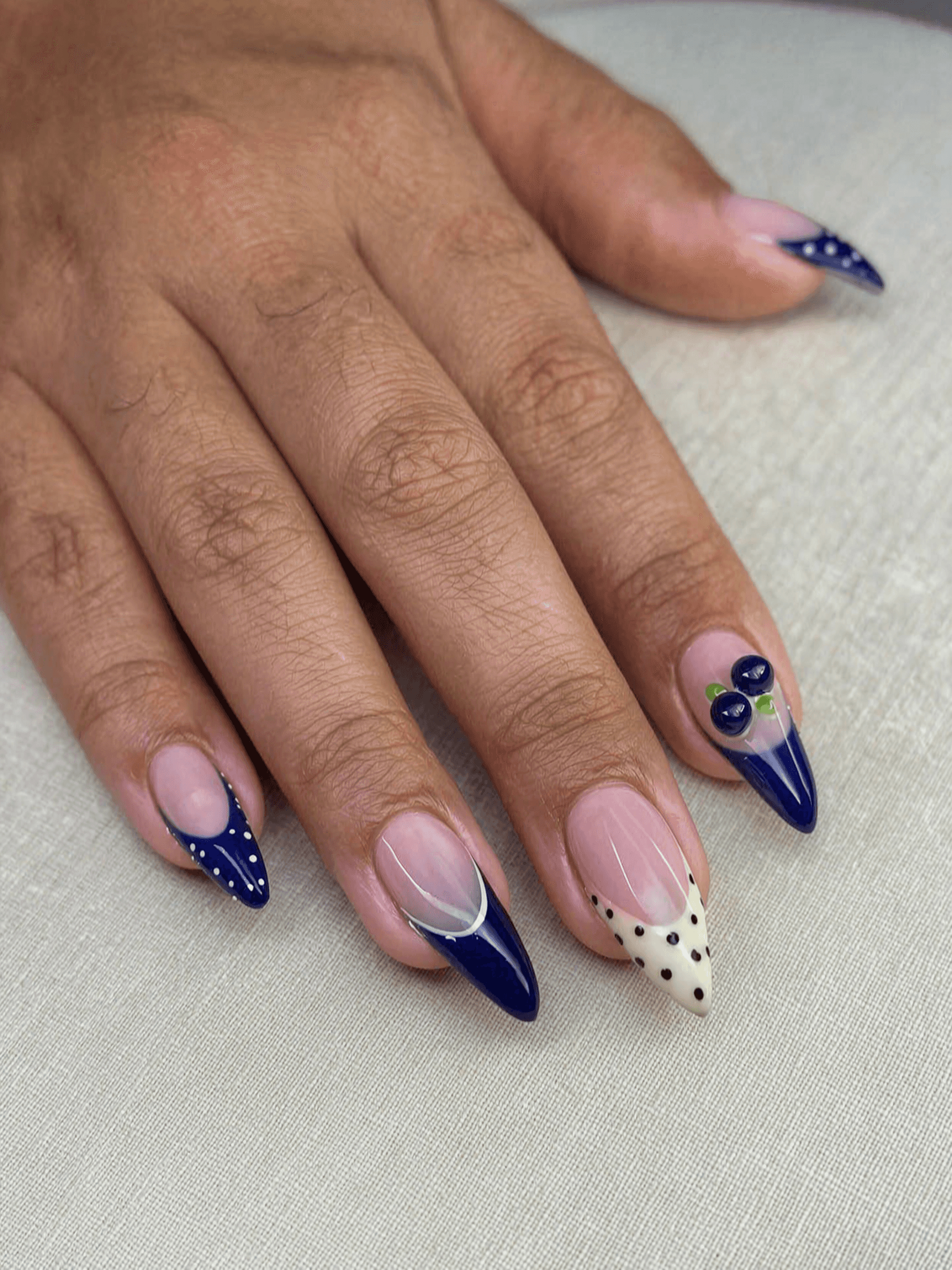 Almond nails with a sheer nude base featuring a mix of navy blue and cream French tips with polka dots, abstract lines, and 3D blueberry art.
