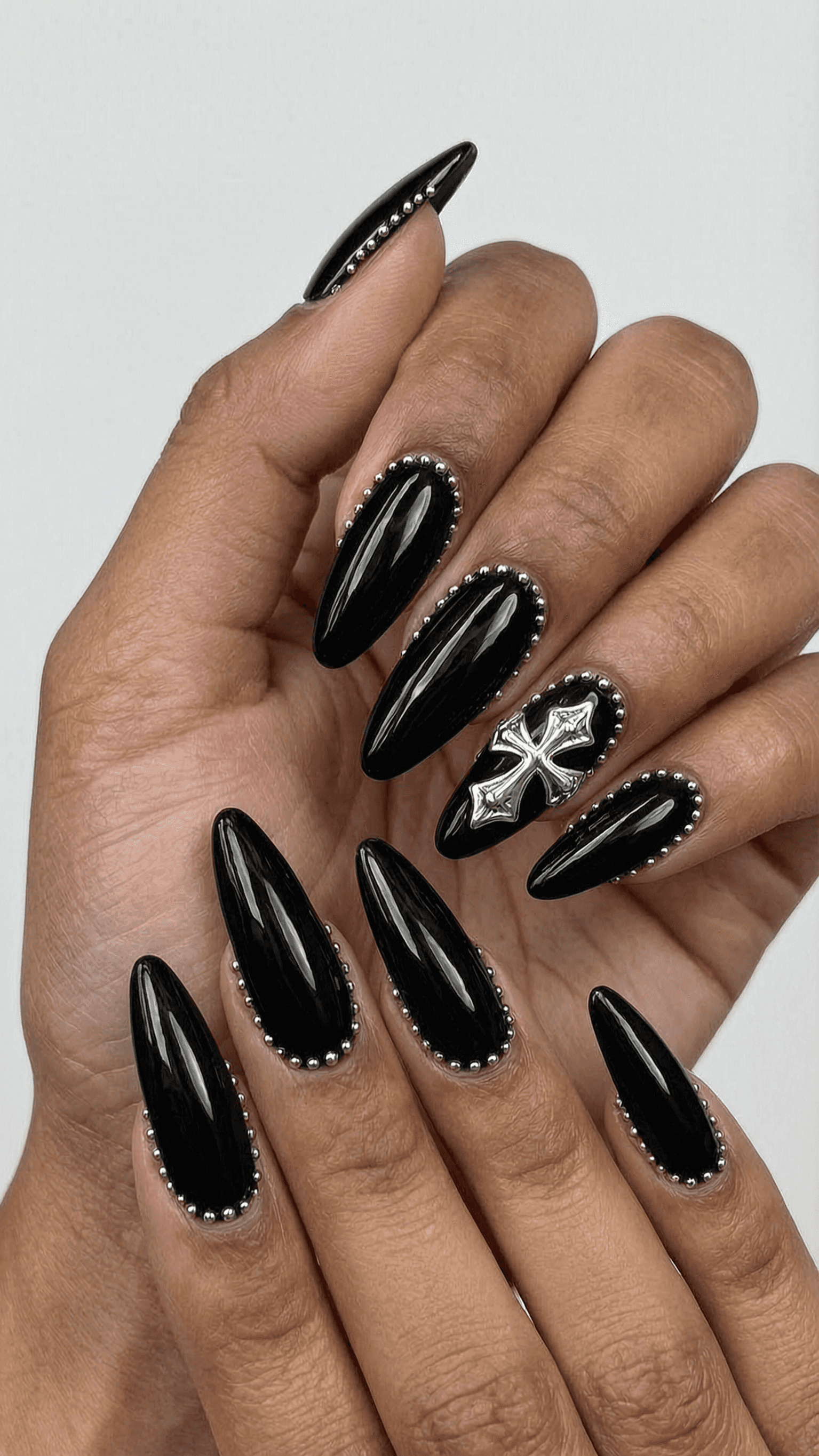 Long black stiletto nails with silver studs and cross charm embellishments, glossy finish.