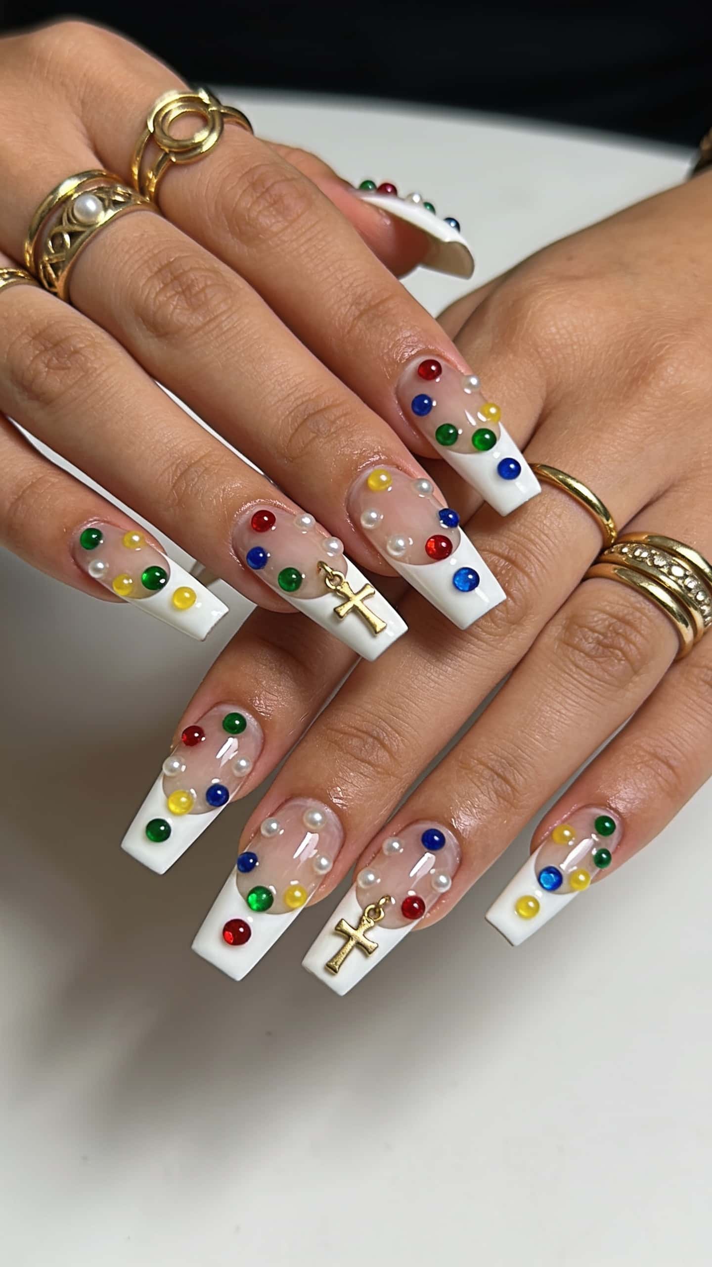 Long square nails with white French tips, adorned with colorful gems, pearls, and a gold cross charm.