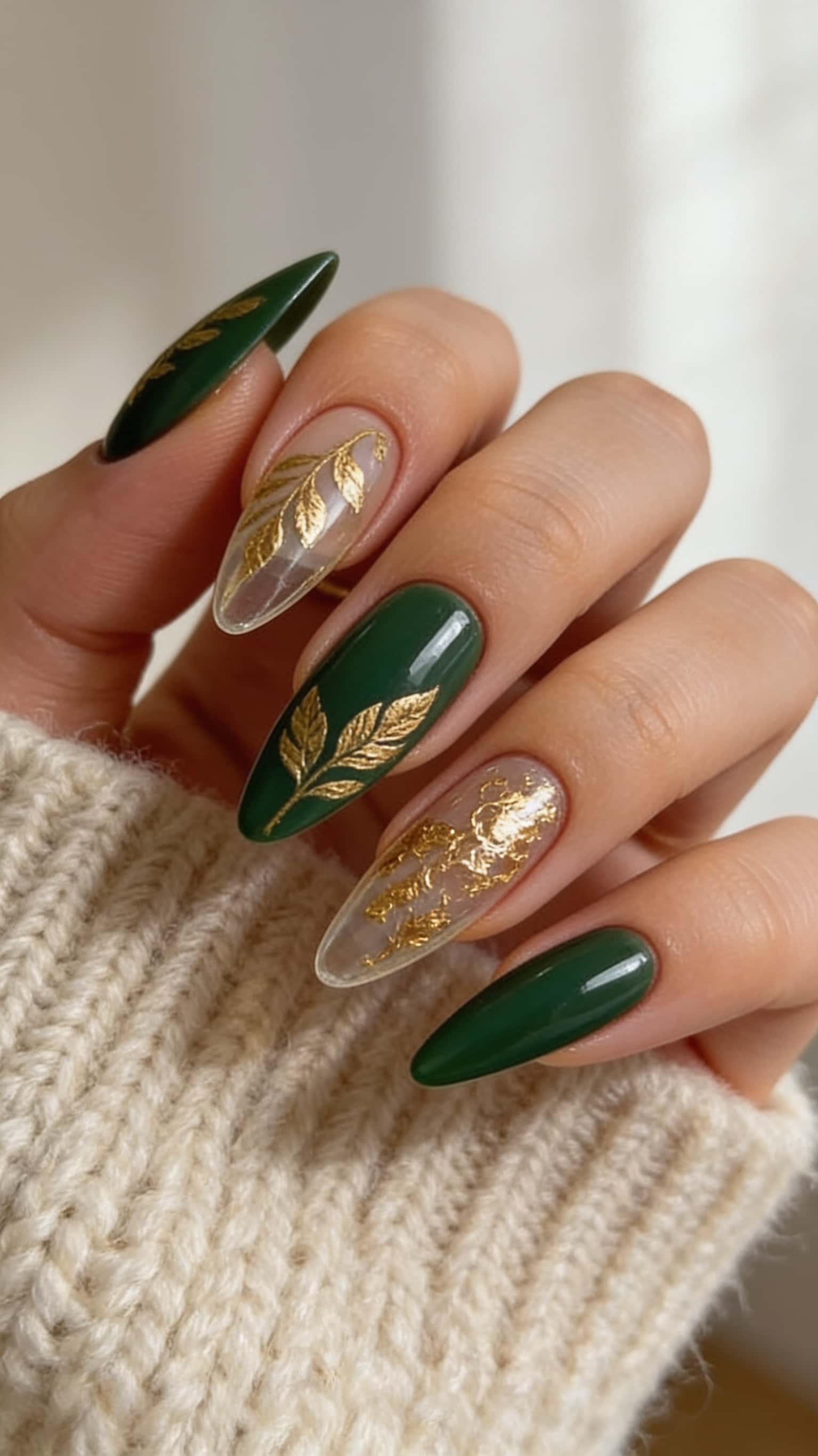 Long almond nails with alternating dark emerald green and clear bases, embellished with elegant gold leaf designs.