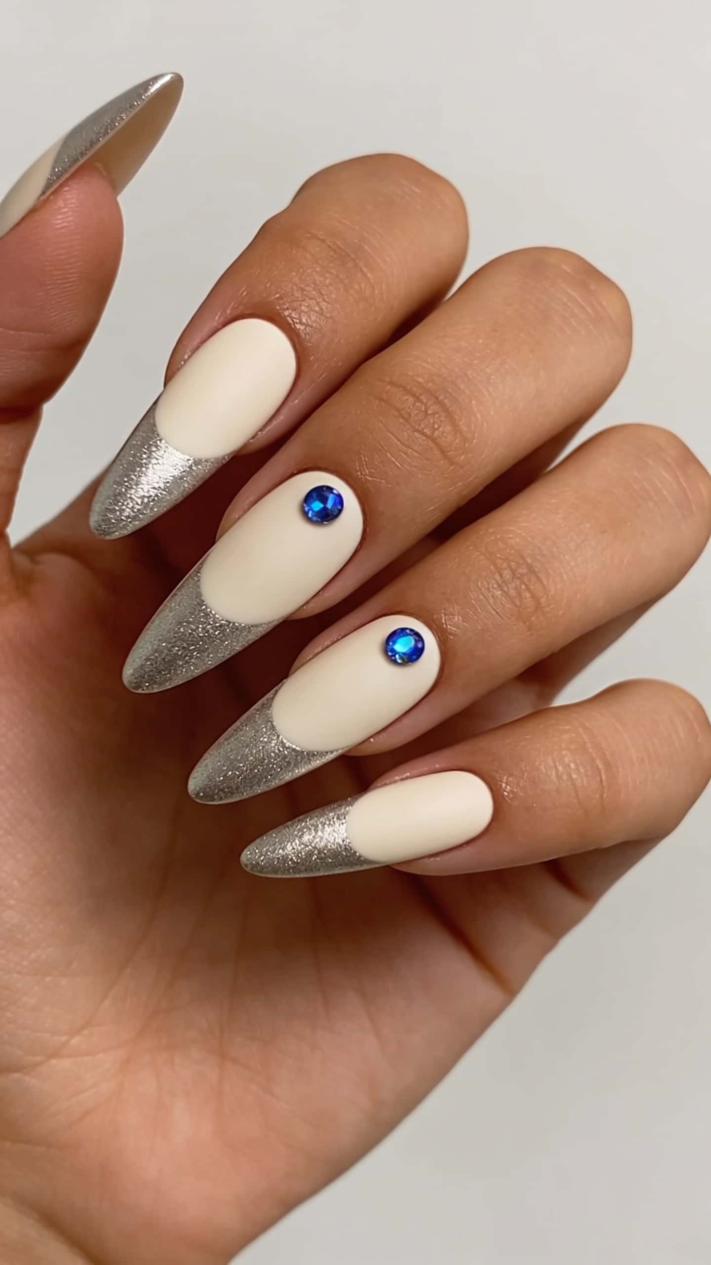 Long stiletto nails with matte cream base, silver glitter French tips, and blue rhinestone accents.