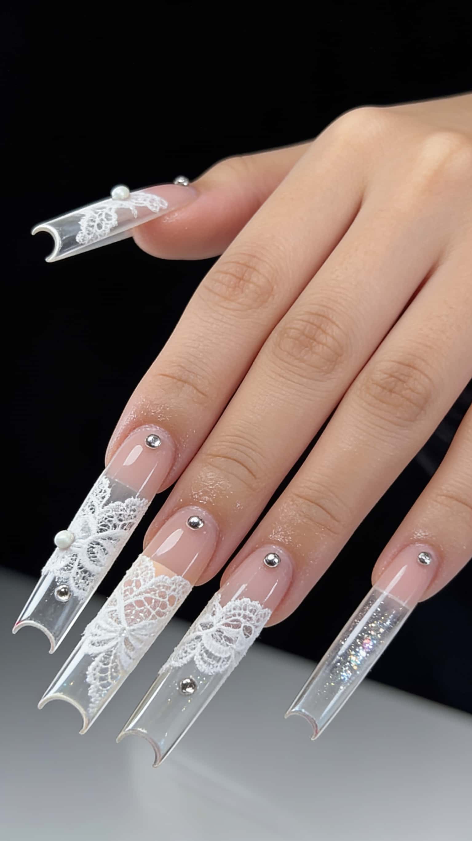 Extra-long pipe nails with a nude base, clear tips featuring white lace patterns, pearls, silver studs, and a glitter accent.