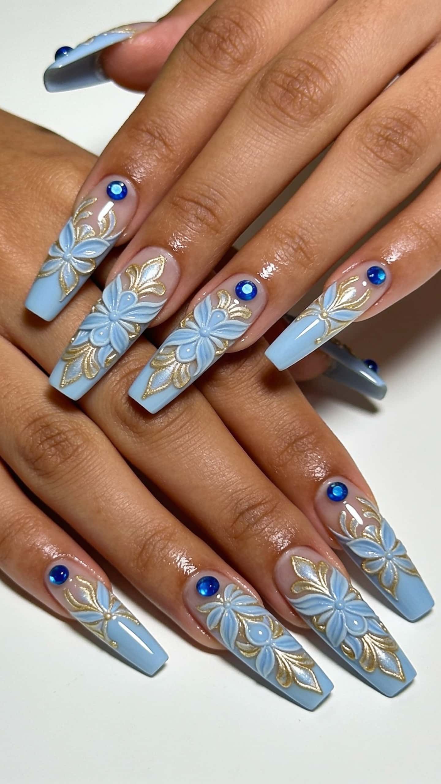 Long coffin nails featuring intricate 3D light blue floral designs with gold accents and blue rhinestones on a clear-to-blue ombre base.