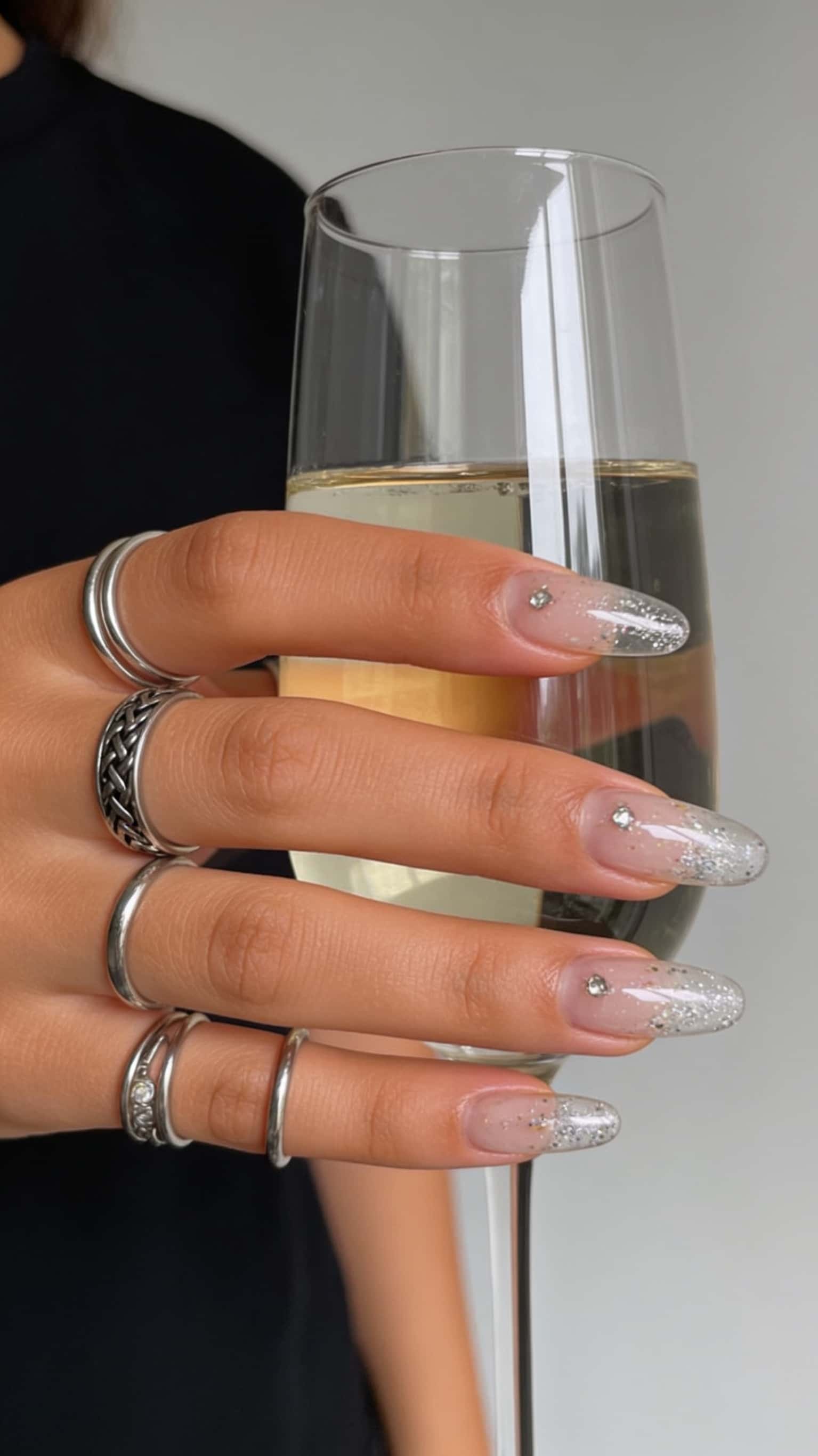 Long oval nails with a sheer nude base, silver glitter ombre tips, and small rhinestone accents.