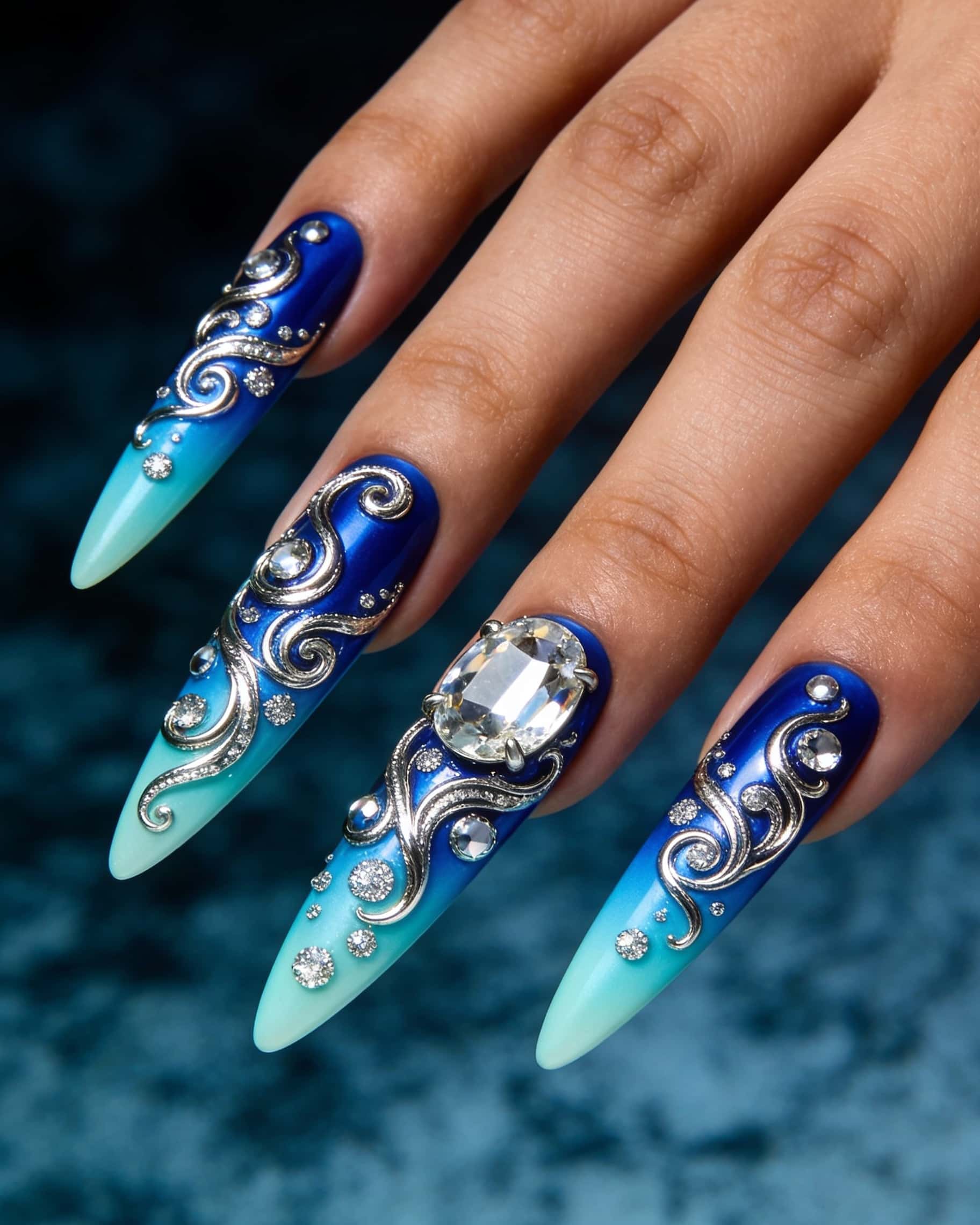 Long stiletto nails with a royal blue base fading into a lighter blue/teal, heavily adorned with intricate silver 3D swirls, large clear rhinestones, and.