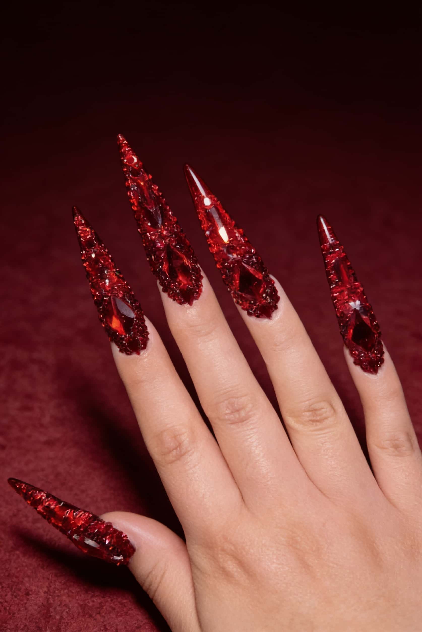 Long stiletto nails fully covered in deep red rhinestones and larger ruby-like gemstones, creating a dazzling, opulent, and dramatic effect