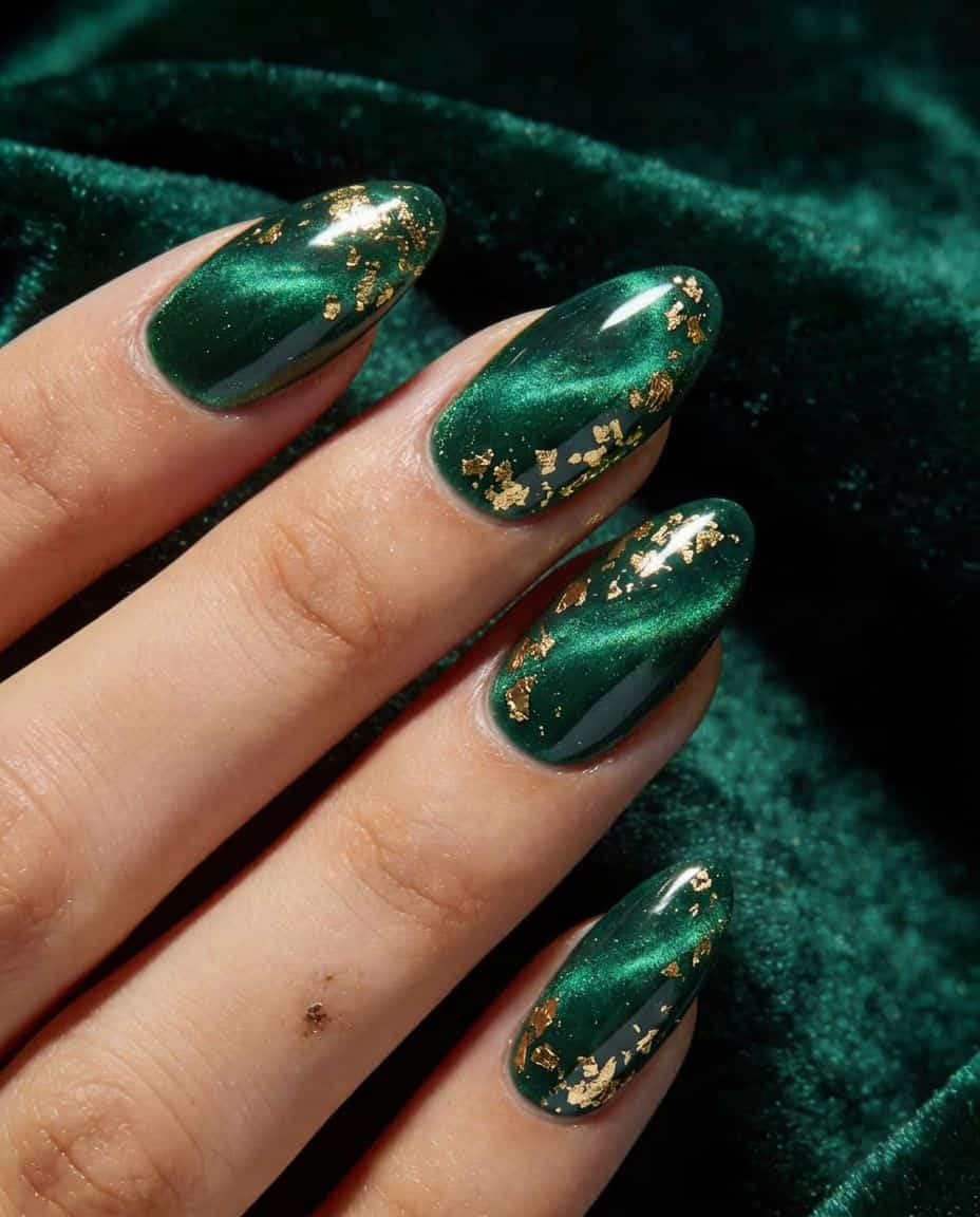 Close-up of a hand with luxurious deep emerald green cat-eye nails, accented with gold foil flakes, resting on dark green velvet.