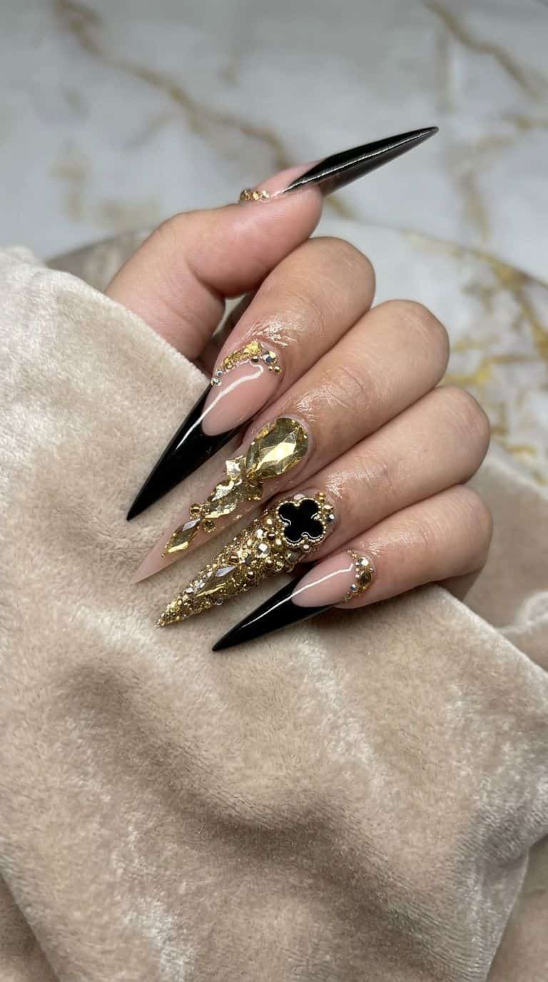 After: Long stiletto nails with nude, black French tips, gold glitter, rhinestones, and a black clover charm.