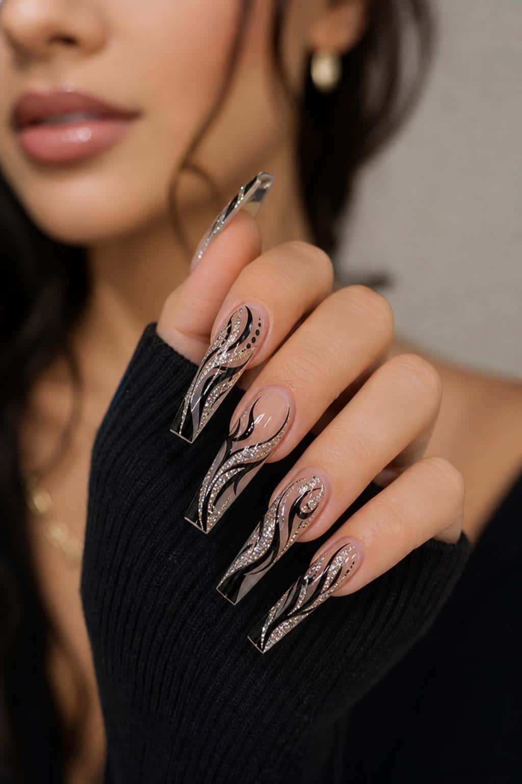 Long coffin nails with a sheer nude base, black abstract swirls, and silver glitter accents.
