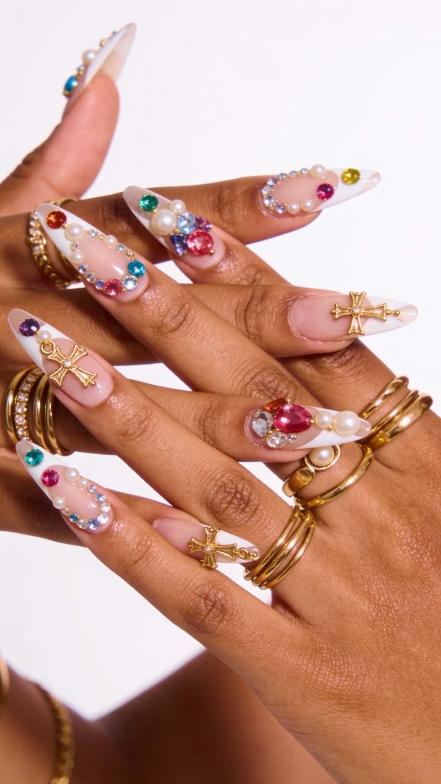 Long stiletto nails with nude and white tips, adorned with colorful jewels, pearls, and gold cross charms.