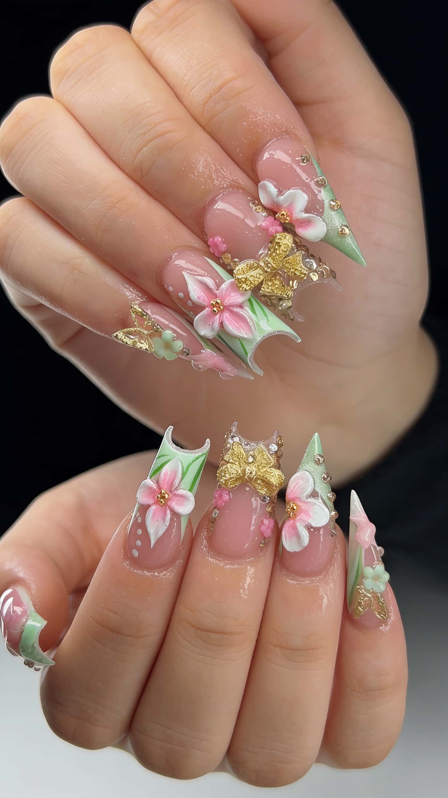 Long stiletto nails adorned with 3D pastel flowers, gold bows, rhinestones, and striped tips on a nude base.
