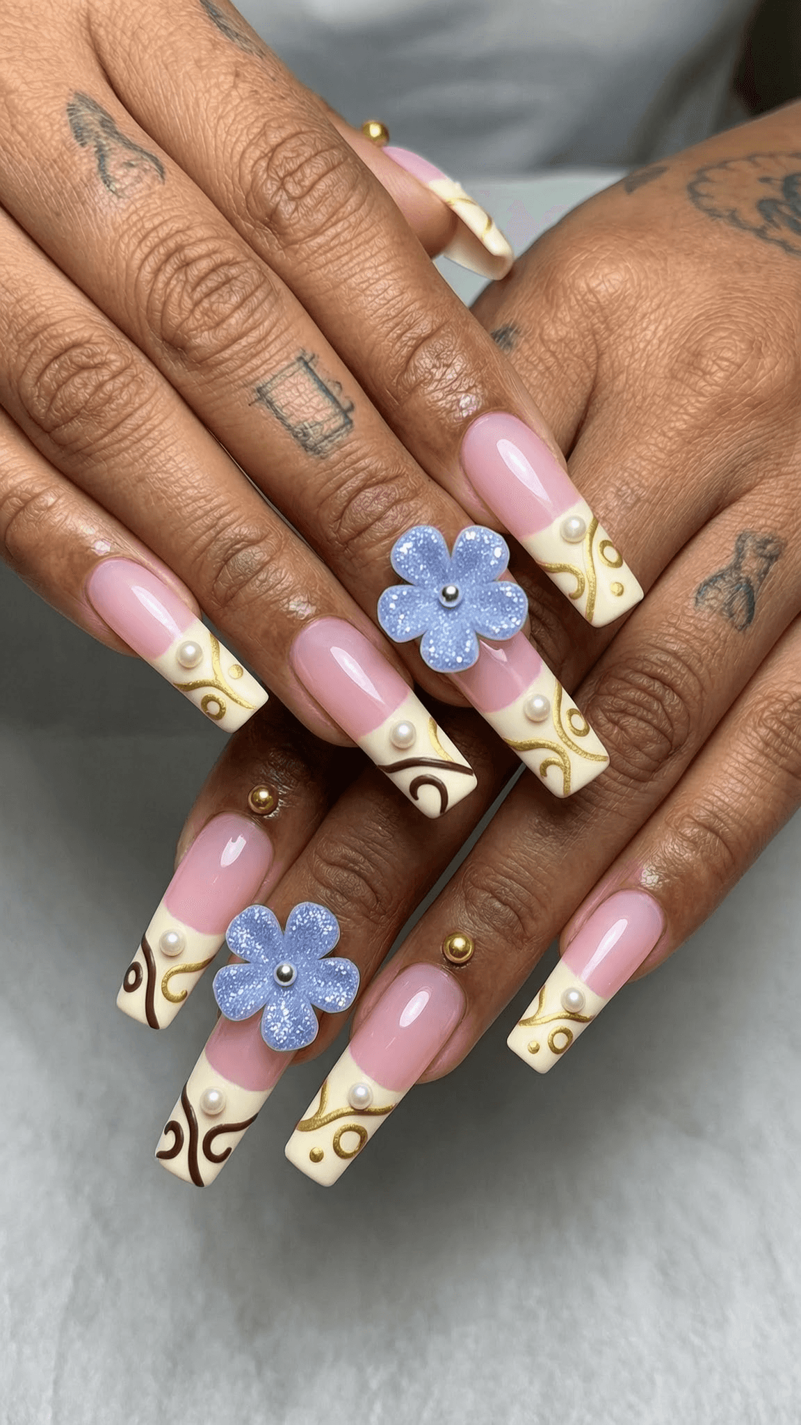 Long coffin nails with pink nude and cream French tips, featuring gold and brown swirl patterns, pearl accents, gold studs, and 3D light blue glitter flowers.