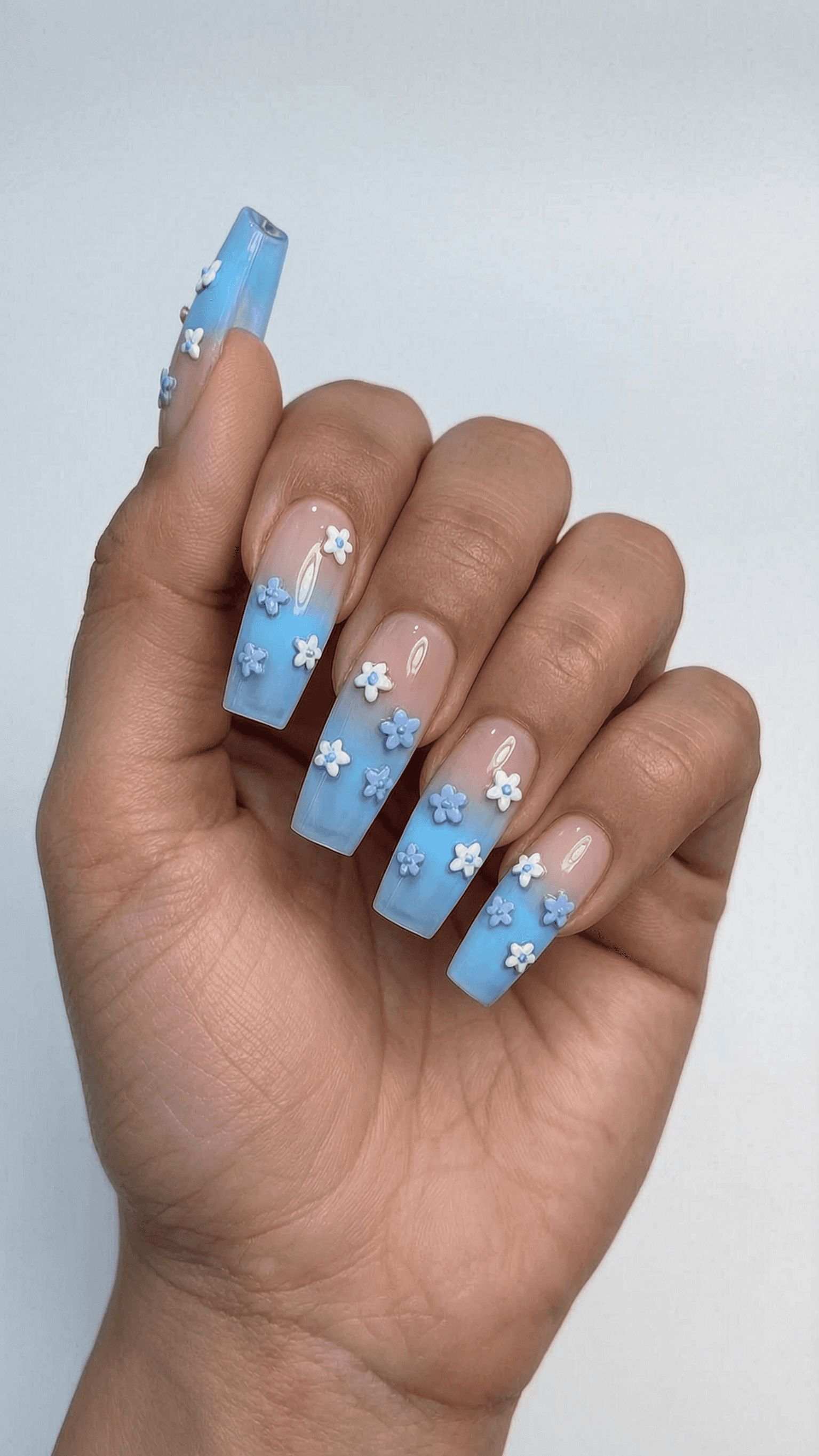 Long coffin nails with a nude to pastel blue ombre, embellished with 3D white and light purple flowers.