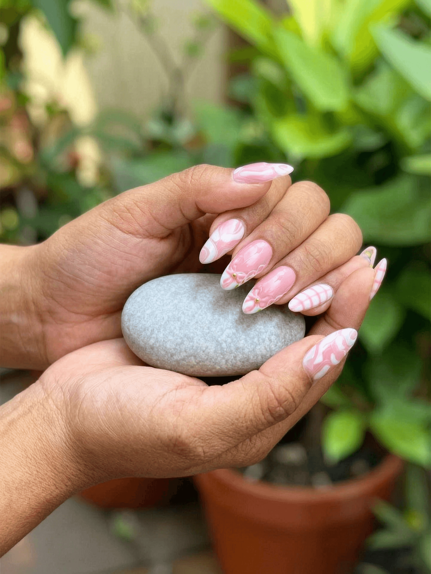 Almond nails with a pastel pink base featuring a mix of white abstract swirls, stripes, and a floral accent with a gold bead.