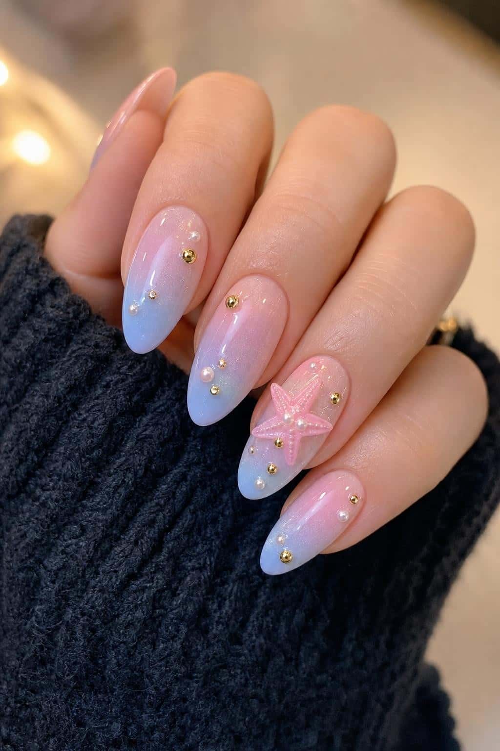 Almond nails feature a pastel pink to blue ombre with shimmer, accented by gold studs, pearls, and a 3D pink starfish.