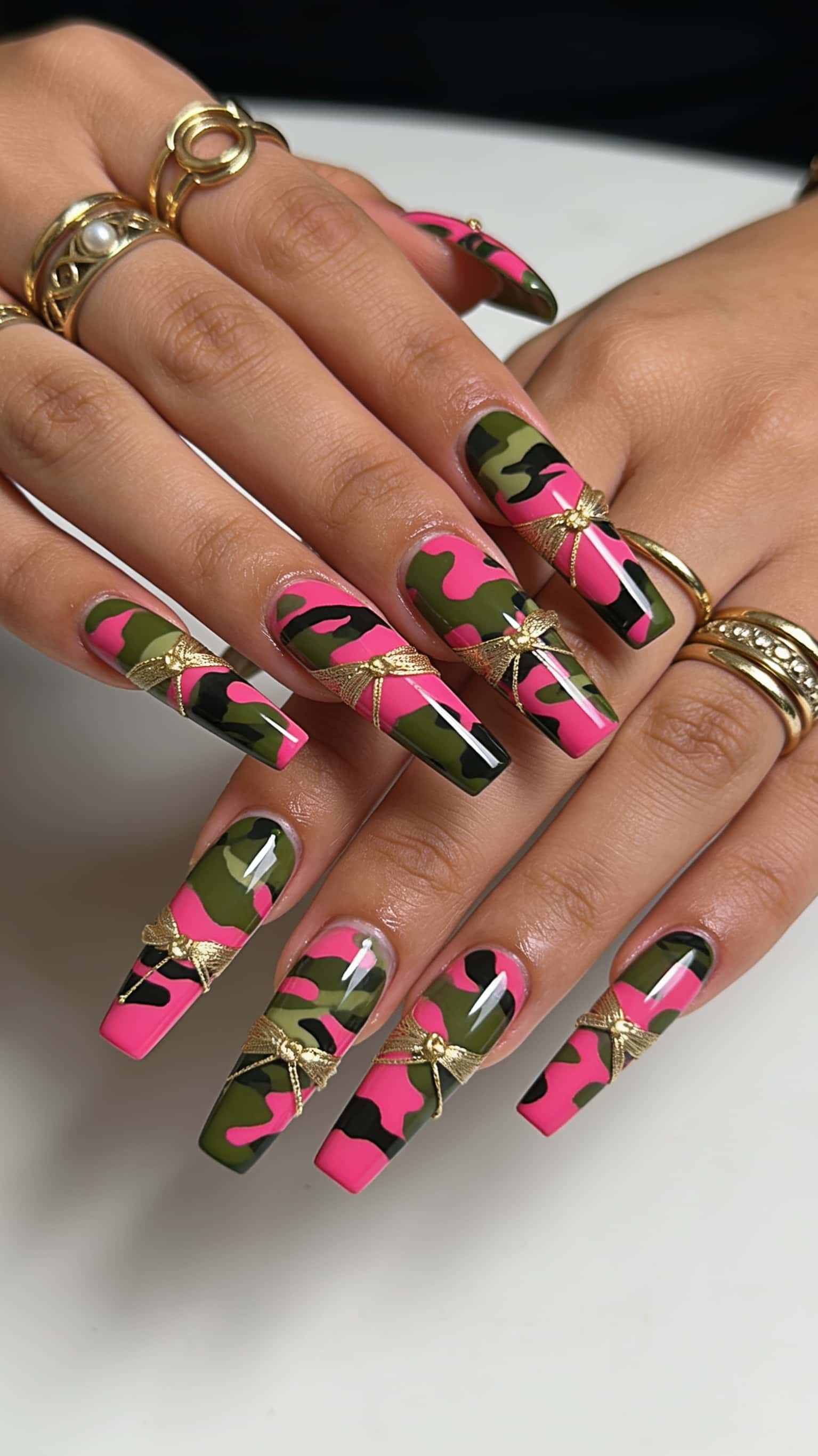 Long coffin nails with a pink, green, and black camouflage pattern, accented by gold ribbon bows.