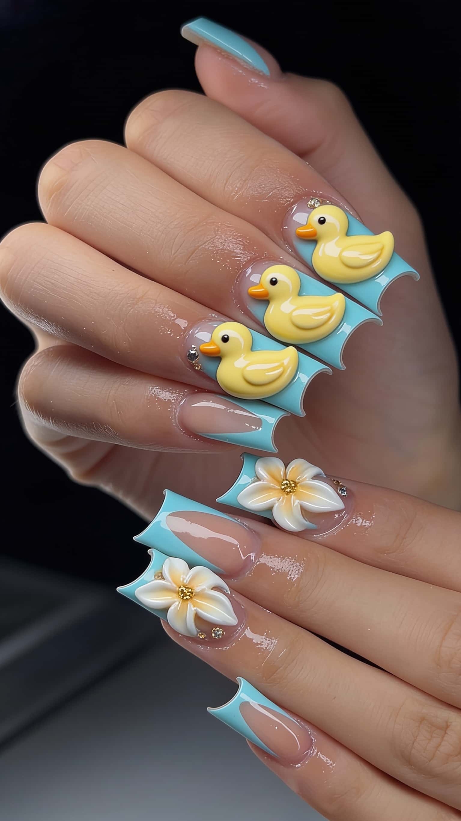 Long flare-tip nails with a nude base and baby blue French tips, adorned with 3D rubber duck and plumeria flower charms, accented with rhinestones.
