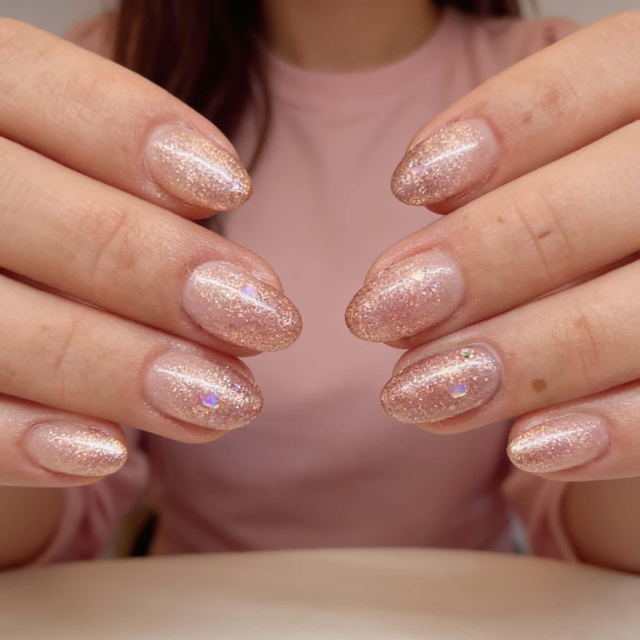 Almond nails with a sheer nude base, full rose gold glitter coverage, high-gloss finish, and small iridescent gems on some nails.