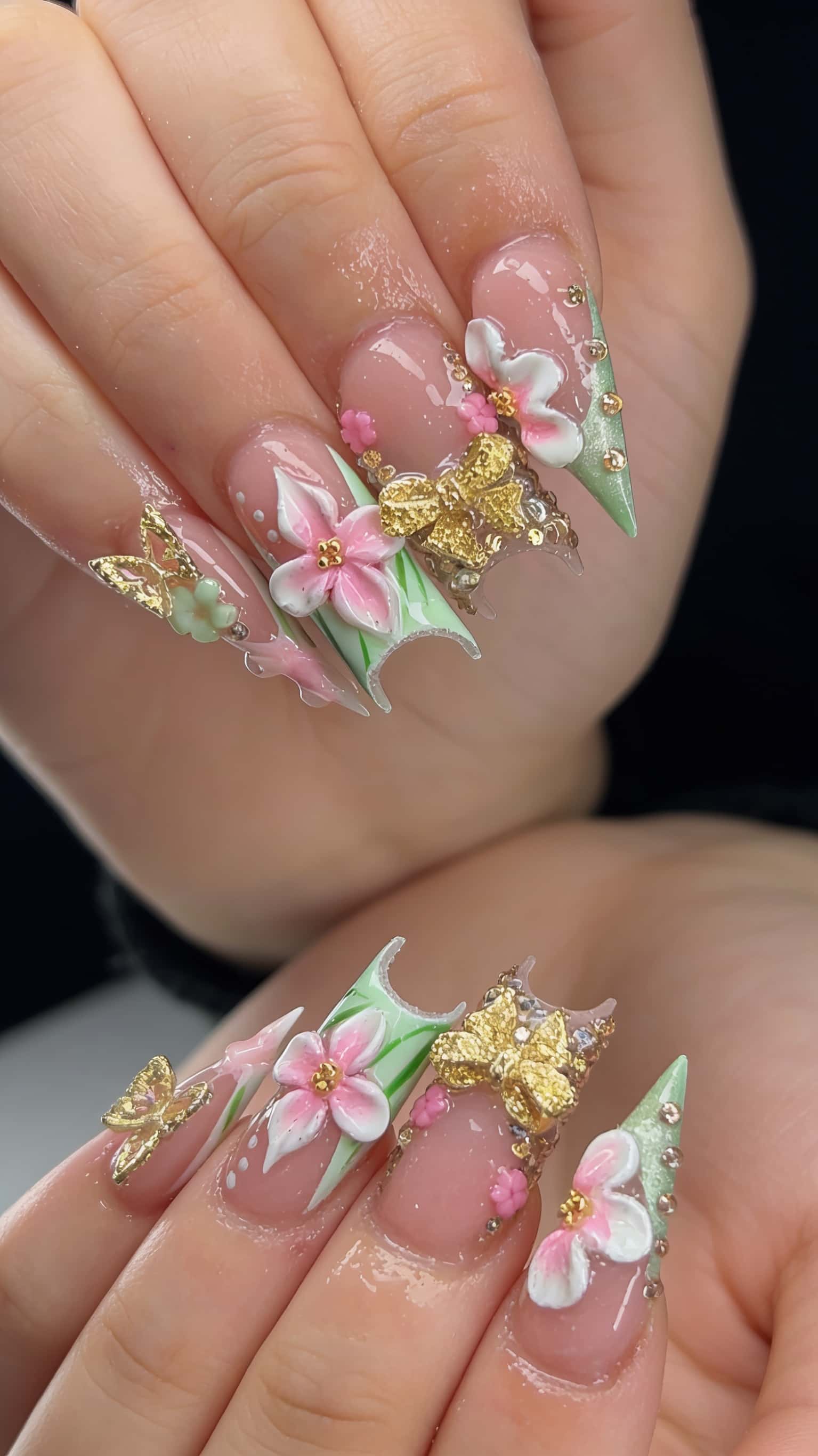 Long stiletto nails with a sheer pink base adorned with 3D sculpted flowers, gold butterflies, rhinestones, and unique green abstract tips.