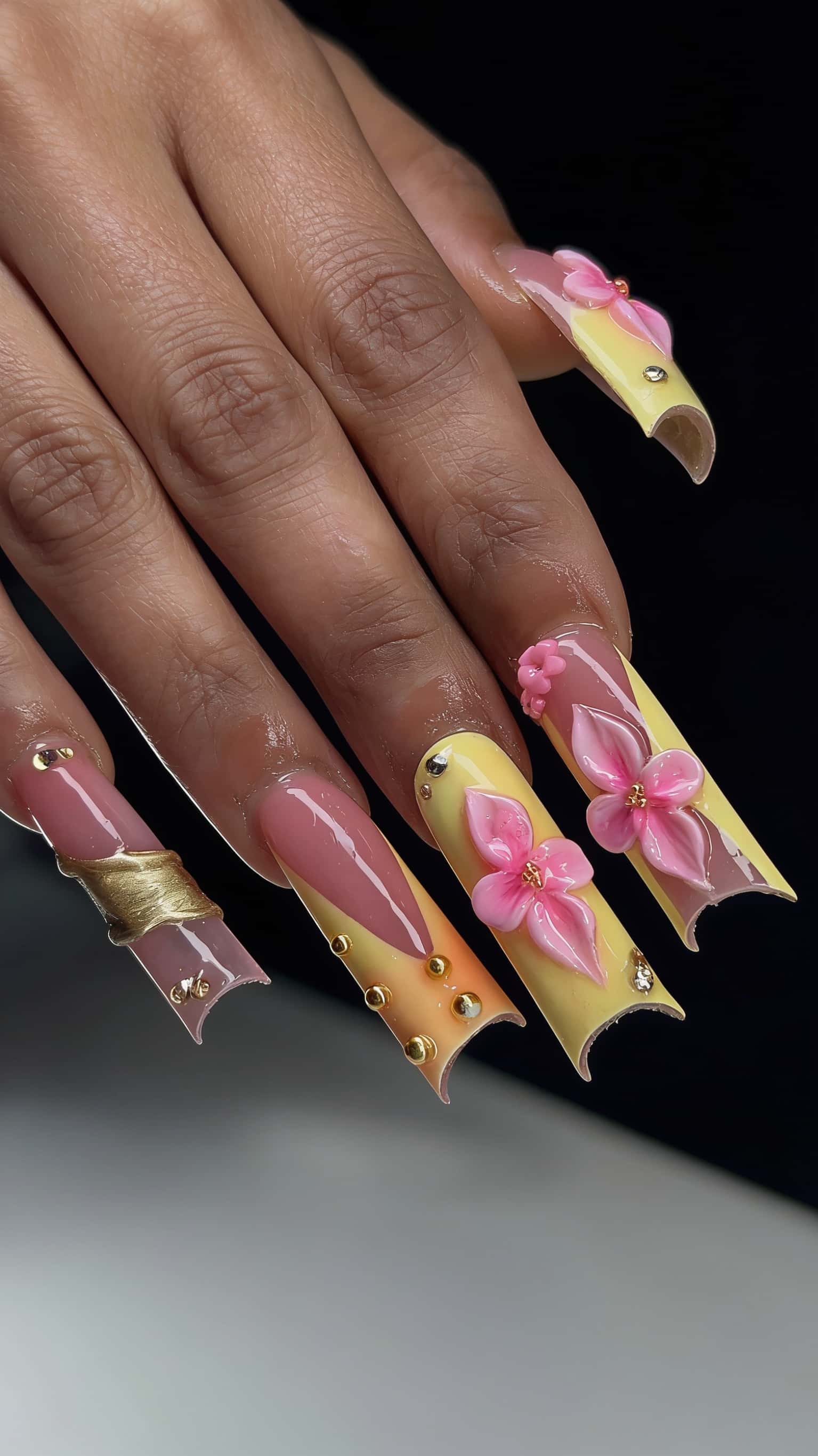 Long, extreme square nails with a pink, yellow, and orange ombre, featuring 3D pink flowers, gold studs, rhinestones, and a sculpted gold accent.