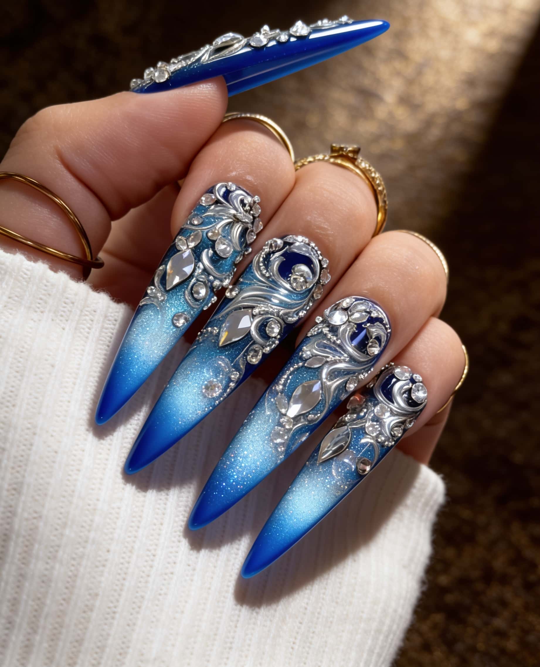 Extra-long stiletto nails featuring a deep royal blue base at the tips, transitioning to a lighter, glittery blue towards the cuticle.